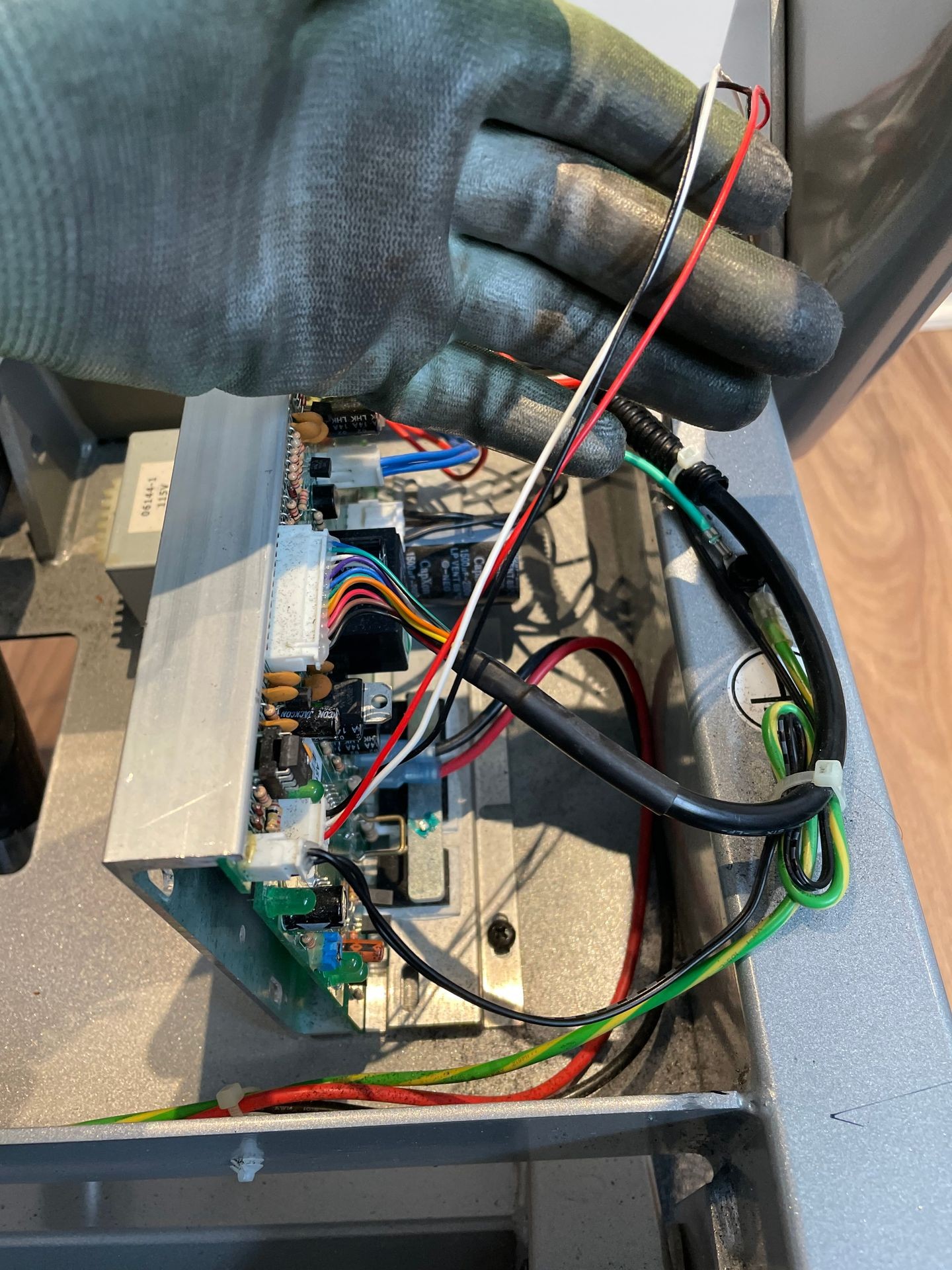 Hand in gray glove holding wires above an open electronic circuit board with colorful cables.